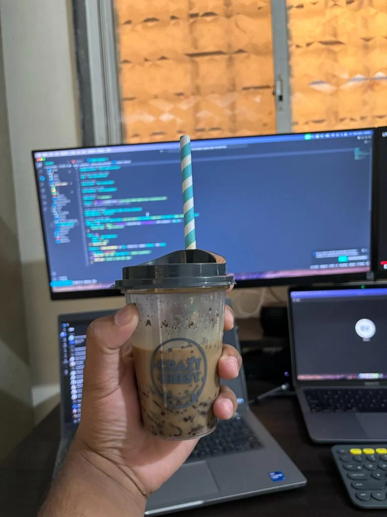 A hand holding a cold drink with computer screens softly blurred in the background.